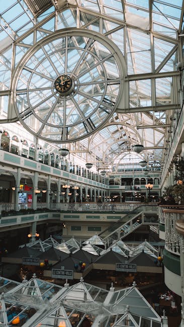 A large, intricate glass-domed ceiling with a prominent circular clock at its center, situated inside Liberty Shopping Centre. The interior features multiple levels with balconies and shops, illuminat