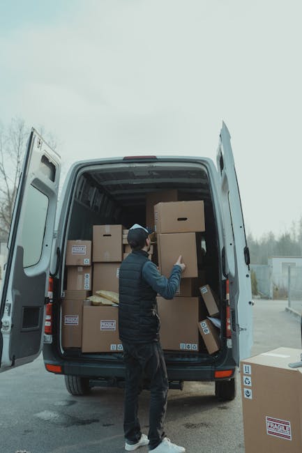 A person wearing a navy blue cap, black quilted vest, and dark pants is inside an open van during a house relocation, surrounded by numerous cardboard boxes of various sizes; some stacked on top of each other and others arranged on the floor of the vehicle. The individual is in the process of unloading or loading the boxes, using their right hand to lift a medium-sized box while facing the rear of the van. The van's interior is visible, showing a spacious cargo area with more boxes arranged neatly. Outside, the scene is set in a paved area with a cloudy sky overhead, and a few trees and structures are visible in the background. The moving process appears organized, with the boxes likely prepared for a furniture transport or home relocation, emphasizing the logistical aspects of packing and loading in a professional removal service, such as provided by Romford Man and Van.