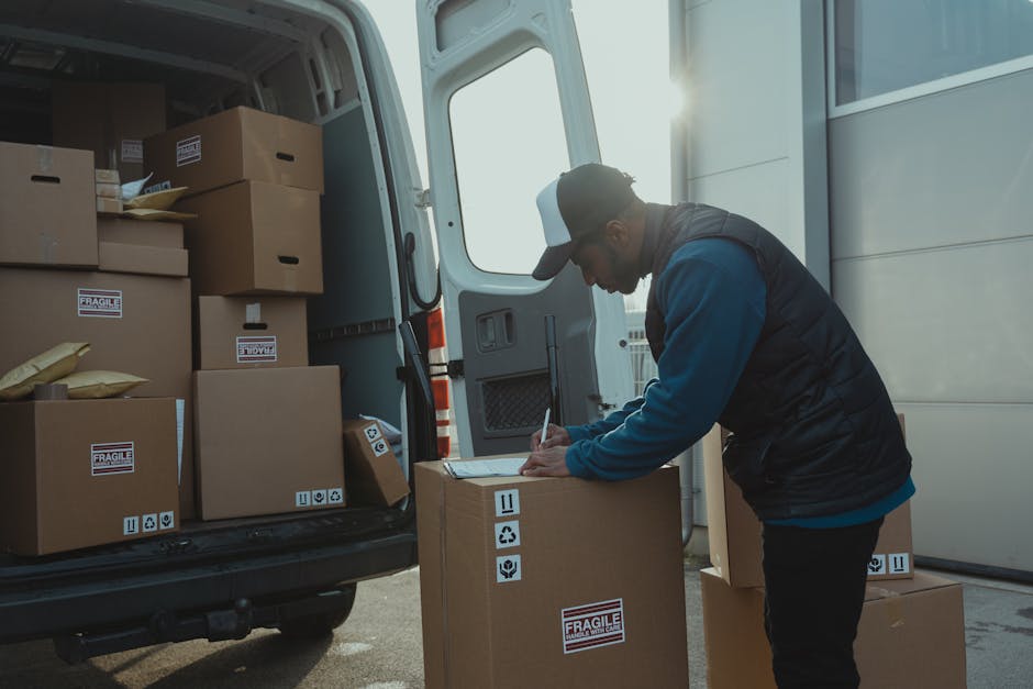 A man wearing a dark baseball cap, a blue long-sleeve shirt, and a black vest is standing outside near the back of a white van, which is parked on a driveway or street during daylight. The open van door reveals a cargo area filled with numerous cardboard boxes of various sizes, some labeled with 'FRAGILE' stickers and packing symbols such as 'this side up' and 'handle with care.' The man is engaged in a home relocation process, busy writing on a clipboard that rests on one of the boxes positioned on the ground, likely verifying inventory or packing details. The van's interior contains additional boxes stacked neatly, some sealed with packing tape, with a few items wrapped in plastic or paper for protection during transport. The scene captures part of a furniture transport or packing and moving operation, emphasizing careful documentation and organized loading practices essential for efficient house removals. Romford Man and Van is involved in the logistics of furniture transport and home relocation services, ensuring smooth handling of household items during moving jobs.