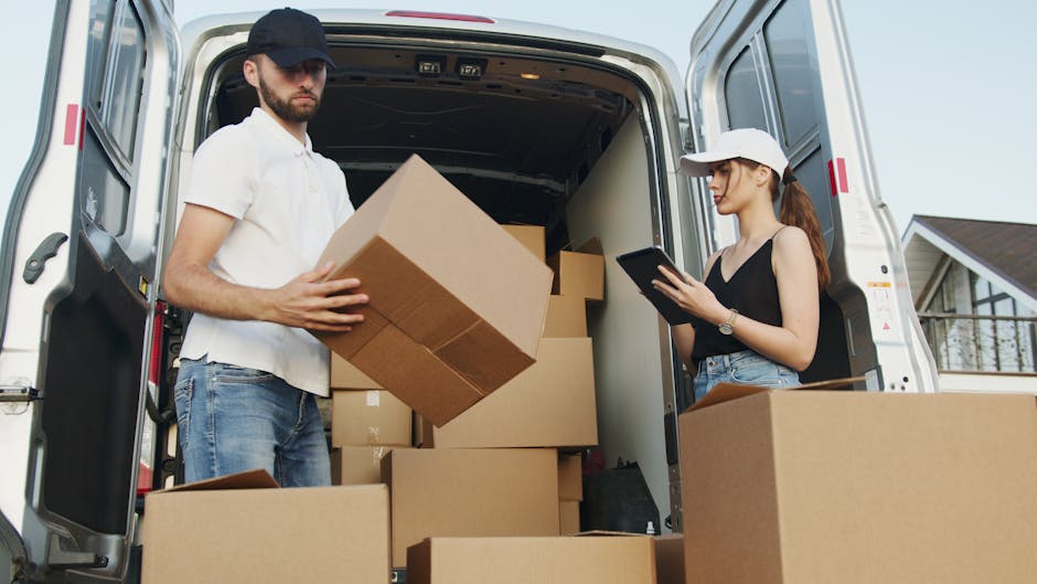 A person wearing a navy blue cap, black quilted vest, and dark pants is inside an open van during a house relocation, surrounded by numerous cardboard boxes of various sizes; some stacked on top of each other and others arranged on the floor of the vehicle. The individual is in the process of unloading or loading the boxes, using their right hand to lift a medium-sized box while facing the rear of the van. The van's interior is visible, showing a spacious cargo area with more boxes arranged neatly. Outside, the scene is set in a paved area with a cloudy sky overhead, and a few trees and structures are visible in the background. The moving process appears organized, with the boxes likely prepared for a furniture transport or home relocation, emphasizing the logistical aspects of packing and loading in a professional removal service, such as provided by Romford Man and Van.